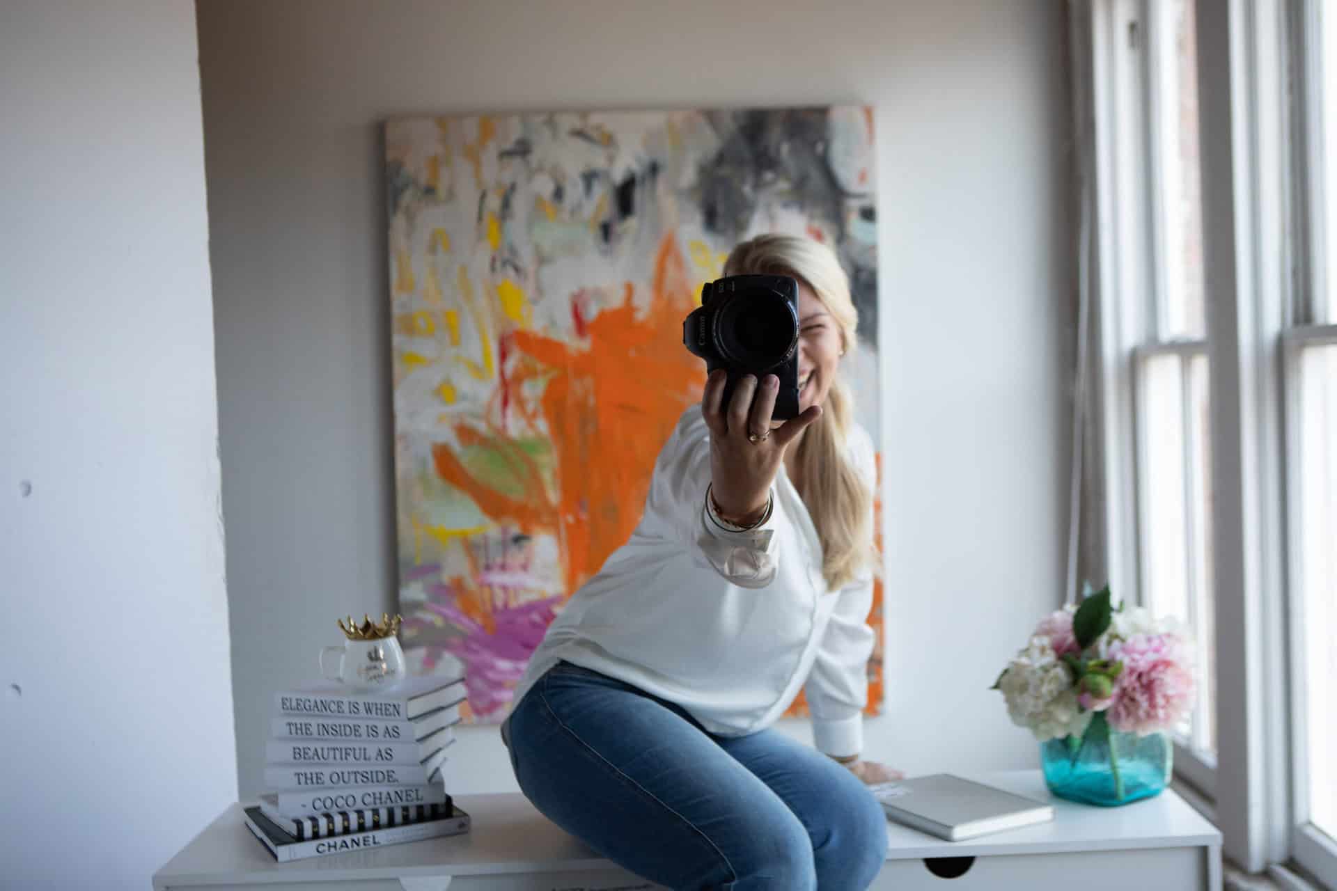 Woman taking a photo with a camera, sitting in front of a mirror in a room decorated with a colorful painting and flowers.