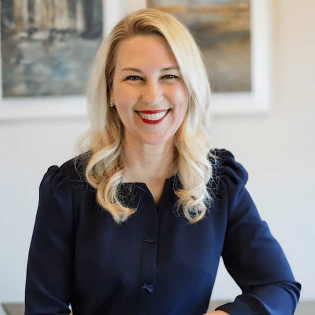 A smiling woman with blond hair, wearing a navy blouse, standing in front of a white wall adorned with abstract paintings.