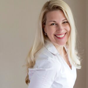 A smiling woman with long blonde hair, wearing a white shirt, standing against a light neutral background.